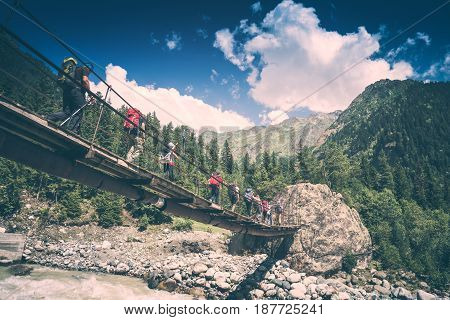 Group of hikers with backpacks go on a suspension bridge over the mountain river. Caucasian mountains Georgia Svaneti region. Instagram stylisation.