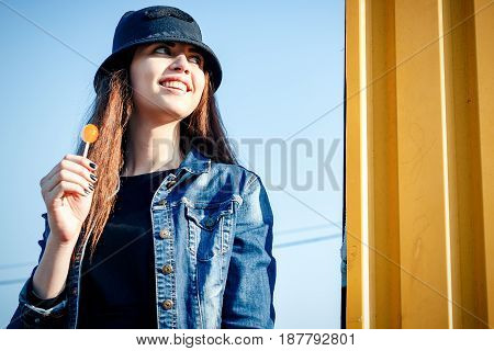 Cheerful Stylish Young Girl In Hat, And Jeans Jacket With Candy On Stick, Chupa Chups, On Sunny Day