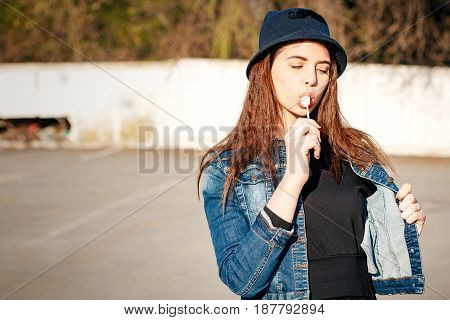 Cheerful Stylish Young Girl In Hat, And Jeans Jacket With Candy On Stick, Chupa Chups, On Sunny Day