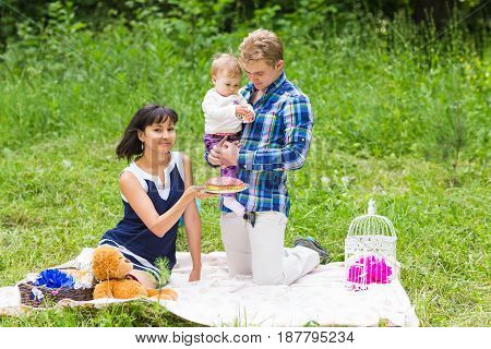 Happy young mother and father with their baby daughter relaxing on a blanket in a park celebrating with birthday cake.