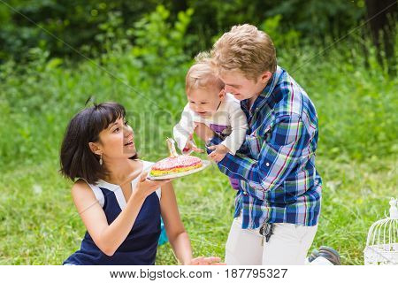 Happy young mother and father with their baby daughter relaxing on a blanket in a park celebrating with birthday cake.
