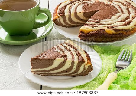 Delicious homemade chocolate cake with cottage cheese Zebra (Marble cake). Tasty breakfast. Piece is cut from cake green cup of tea and green gauze textiles on white wooden table. Selective focus