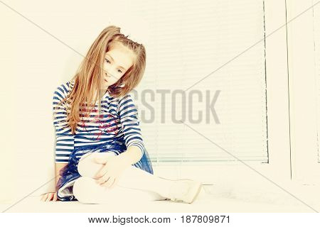 Little girl in blue striped dress and a white bow on her head.She sits at the window with sad eyes something lost in thought.Creative toning of a photograph.