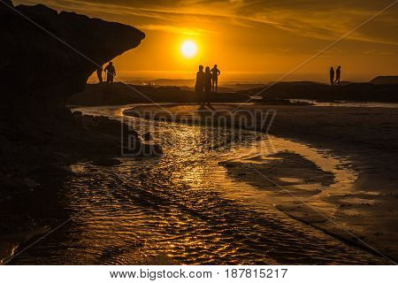 People and rock formations on a beach silhouetted by a golden orange sunset