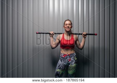 Young sporty smiling woman wearing red top with bodybar on shoulders. Sports girl posing against grey wall. Fitness, sports and healthy lifestyle.