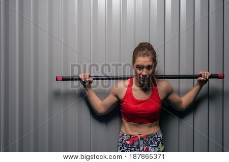 Portrait of young sporty woman wearing red top with bodybar on shoulders. Sports girl posing against grey wall. Fitness, sports and healthy lifestyle.