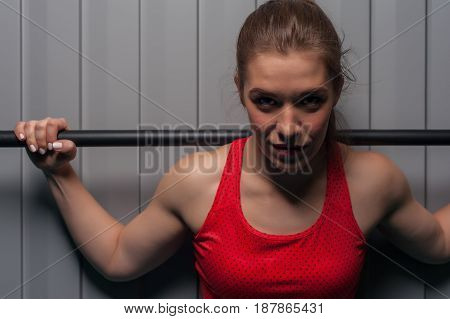 Portrait of young sporty woman wearing red top with bodybar on shoulders. Sports girl posing against grey wall. Fitness, sports and healthy lifestyle.