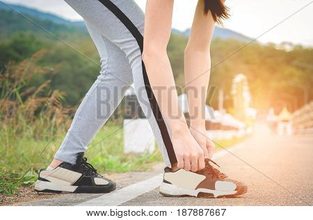 Shot of young woman runner tightening running shoe laces getting ready for jogging exercise outdoors. Female jogger lacing her sneakers standing on road path before morning run.