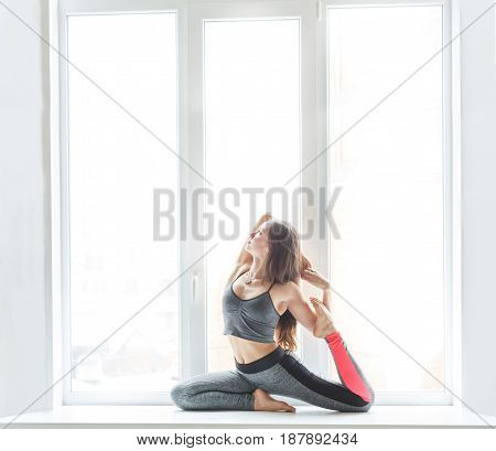 Young woman makes yoga pose at window backlight, copy space. Girl do mermaid posture. Healthy lifestyle