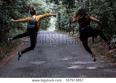Heel click on forest path by young adult girls / women sisters with hair in bun and leather backpack with mustard yellow sweater jumping in the air