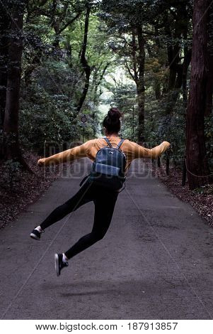Heel click on forest path by young adult girl woman with hair in bun and leather backpack with mustard yellow sweater jumping in the air