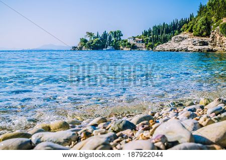 Pebble beach near Kalami with pine and cypress trees and an yacht at anchor in a bay on background. Corfu Island, Greece.
