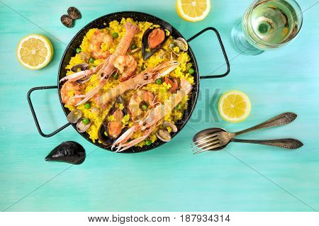 Photo of Spanish seafood paella in typical paellera, with slices of lemons, mussels and clams shells, glass of white wine, fork and spoon, and a place for text, shot from above on a teal texture