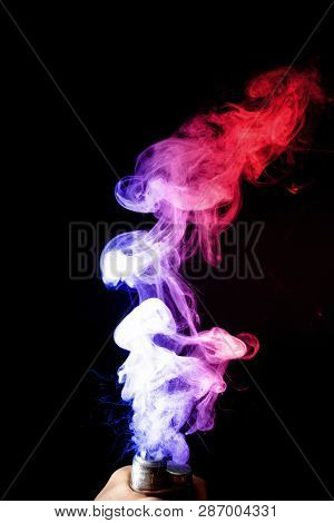 Male Hand Holds Vape. Vape Clouds Splash From Vape Coil On Black Background. Fog Is Dual Color -- Bl