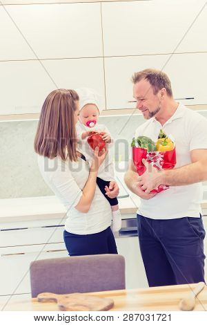 Happy Parents Standing In The Kitchen And Prepares For Cooking With Their Baby Girl Chef. Father Bri