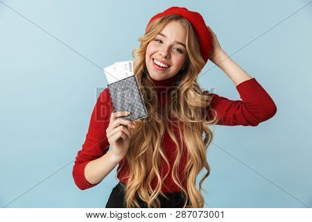 Photo of modest blond woman 20s wearing red beret holding passport and travel ticket isolated over blue background in studio