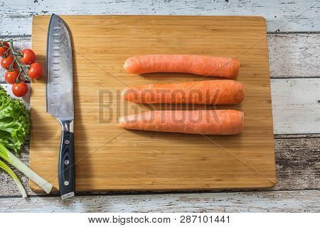 Santoku Kitchen Knife On A Cutting Board With Fresh Vegetables: Carrots, Tomatoes, Lettuce And Green