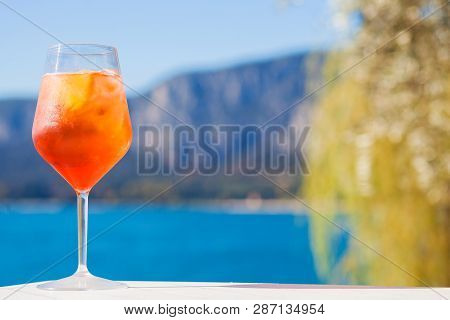 Glass Of Delicious Aperol Spritz Cocktail On Blurred Sunny Lago Di Garda Background
