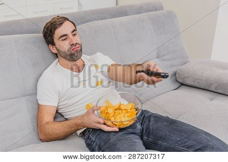 The Guy In The T-shirt Sits On The Couch, Eats Chips And Watches The Tv. While Holding The Remote In
