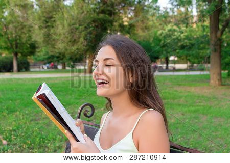 Beautiful Young Brownhair Girl Smells A Fresh Printed Book Sitting On The Bench In The Park