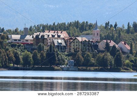 Small Mountain Town With Tall Densely Built Family Houses Around Local Church With Clear Lake In Fro