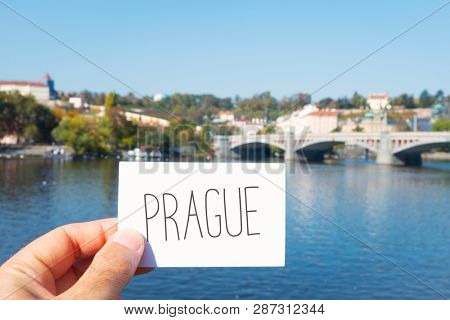 closeup of the hand of a caucasian man with a signboard with the wort Prague, in front of the Vltava River in Prague, Czech Republic, with the Manes bridge and the Prague Castle in the background