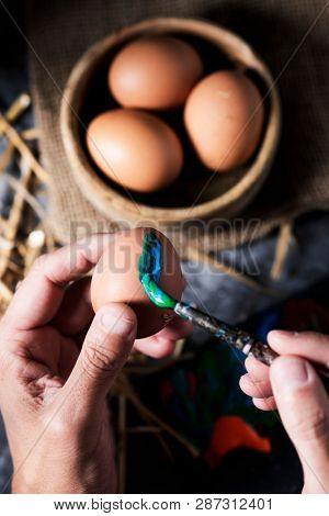 closeup of a young cacausian man painting an easter egg, on a gray rustic wooden table, next to some brown chicken eggs in a boxwood bowl and some straw stalks