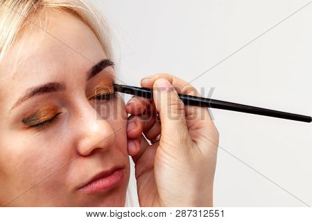 Visagiste With A Brush In One Hand Putting Makeup On The Eyes Of The Model, The Other Hand Pulls The