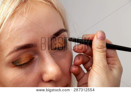 Visagiste With A Brush In One Hand Putting Makeup On The Eyes Of The Model, The Other Hand Pulls The