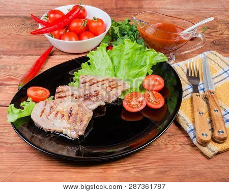Grilled Pork Steaks On Lettuce Leaves And Cherry Tomatoes On Black Dish, Souce, Greens And Vegetable