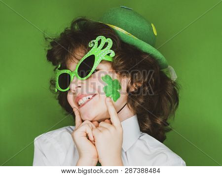 Child Celebrating St. Patricks Day Showing His Make-up. A Small Curly Boy In Green Carnival Accessor