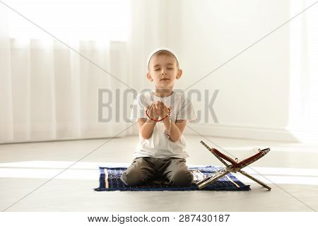 Little Muslim Boy With Misbaha And Koran Praying On Rug Indoors
