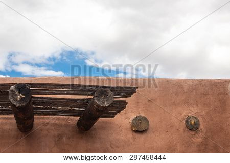 Exterior Detail Of Red Adobe Building With Beams, Patch Of Blue Sky With Clouds, Horizontal Aspect