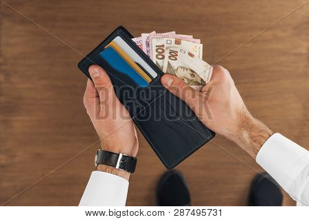 Top View Of Man Puting Hryvnyas Banknotes In Wallet With Credit Cards On Wooden Background