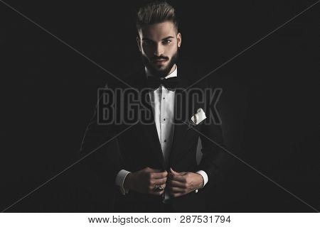 man in tuxedo with bowtie and white hankerchief adjusting his lounge jacket with both hands on black background