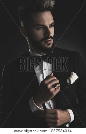 man in tuxedo with bowtie and white hankerchief fixing his sleeve while looking away on black background