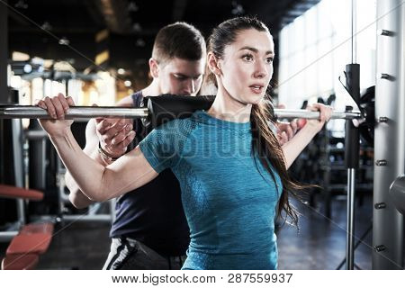 Personal trainer man helps woman work with barbell at squat exercises in gym