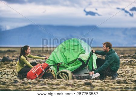 Camping people outdoor lifestyle couple tourists putting up a tent in nature lansdcape at dusk. Campers with backpacks setting up their campsite for the night in mountain summer background.
