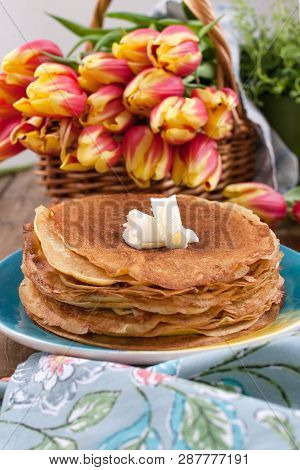 Lots Of Thin Pancakes On A Wooden Background Or Table, And A Basket With Fresh Red-yellow Tulips. Ba