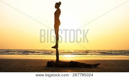 Fit Sporty Couple Practicing Acro Yoga With Partner Together On The Sandy Beach.