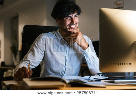Smiling young man working at the office late at night, using computer