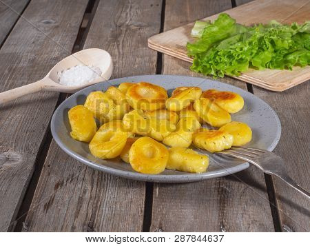 Fried Potato Gnocchina On A Gray Ceramic Plate, Leaves Of Fresh Leaf Lettuce Lie On The Kitchen Boar