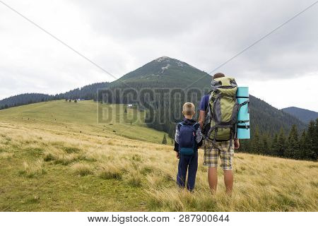Father And Son With Backpacks Hiking Together In Summer Mountains. Back View Of Dad And Child Holdin