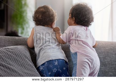 Back view of baby boy and girl standing together on back of couch, cute playing playdate interaction