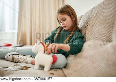 A Small Girl In A Green Sweater And Converses Is Filling Her Unicorn Moneybox With Coins One By One 