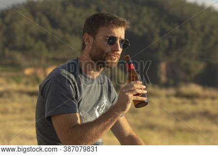 A Young Man, With Glasses And A Beard, Drinks Beer From A Bottle, While Enjoying The Landscape And T