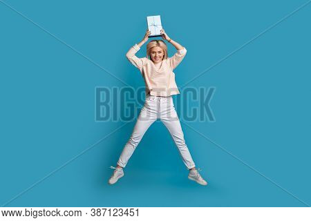 Lovely Blonde Lady Jumping On A Blue Studio Wall Holding Up A Box With Present While Smile At Camera