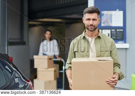 Waist Up Portrait Of Handsome Man Holding Box And Looking At Camera While Standing By Self Storage F