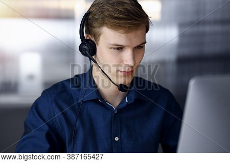 Young Blond Businessman Using Headset And Computer In A Darkened Office, Glare Of Light On The Backg