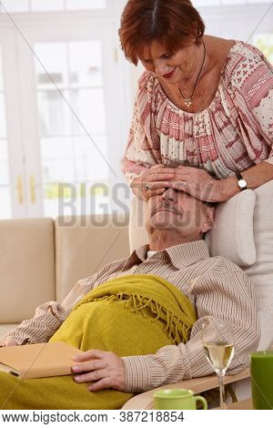 Senior woman surprising her husband resting in armchair in living room.
